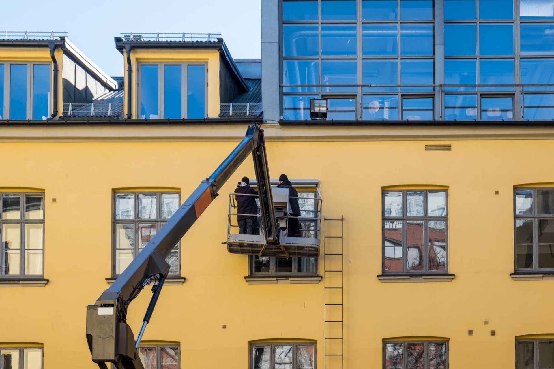 Arbetare i en skylift sysselsatta med att förnya fasaden på en gul byggnad försedd med omfattande fönsterpartier